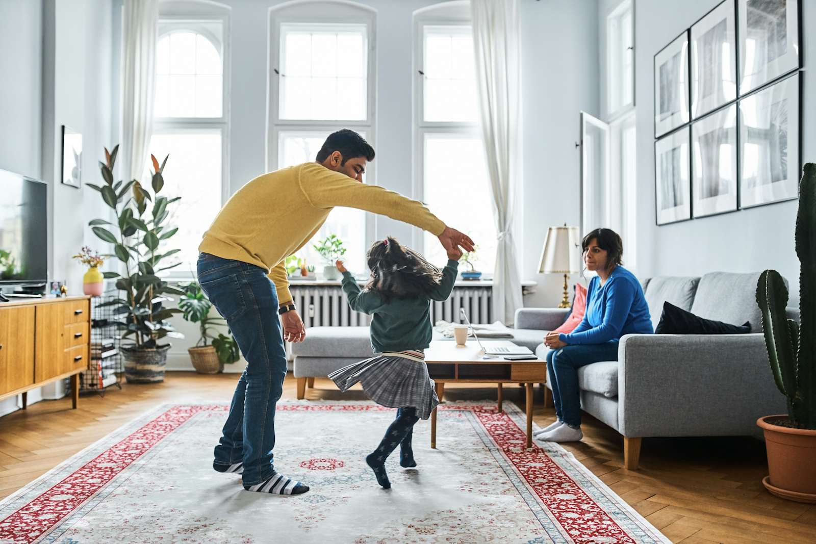 Woman looking at man and daughter while sitting on sofa. Girl is dancing with father in living room.